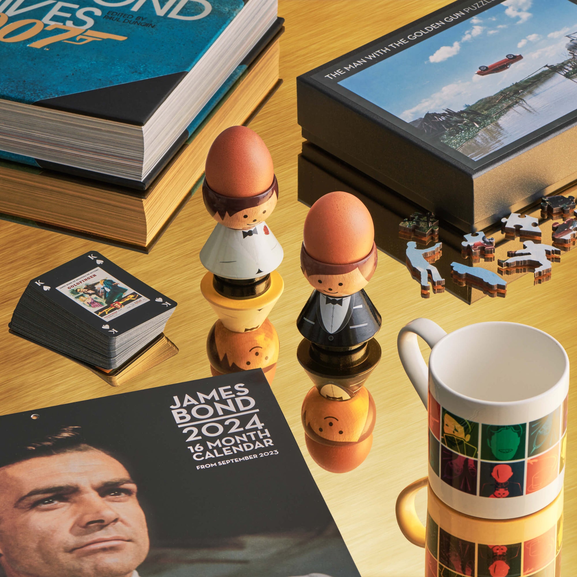 Two kokeshi dolls in tuxedos on a table with James Bond books, a calendar, and a mug.