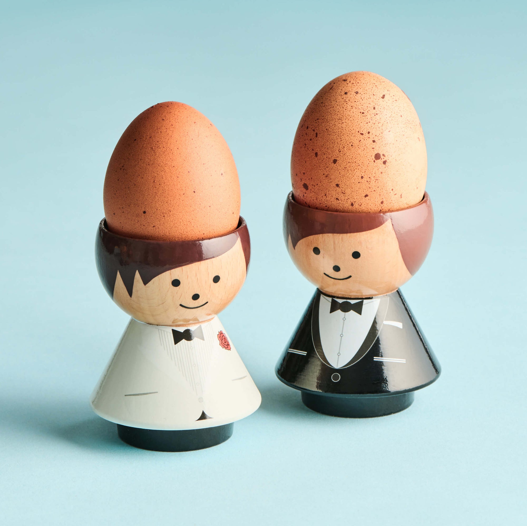 Two kokeshi doll egg cups, one in a white tuxedo and the other in black, each holding a brown egg.