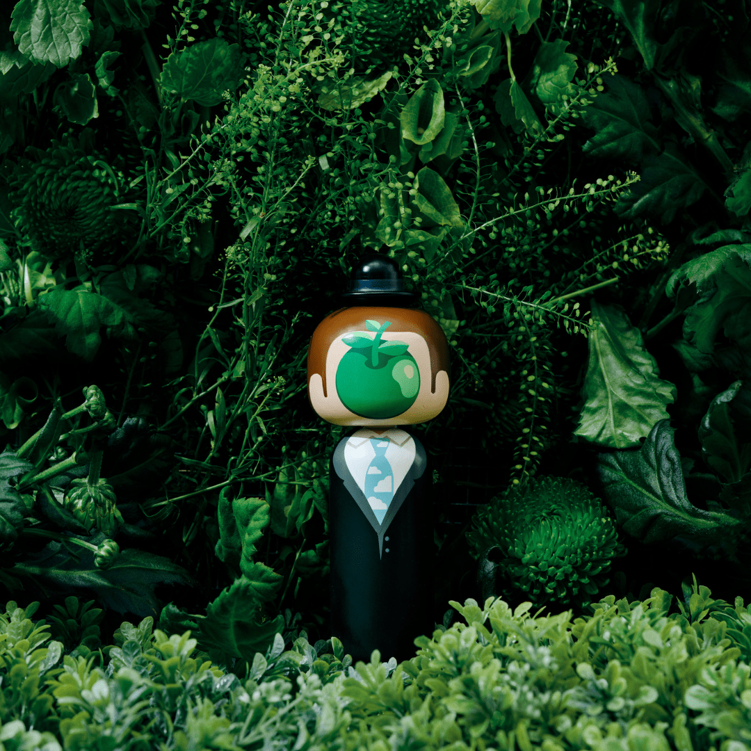 A kokeshi doll with a green apple face stands amidst lush green foliage.