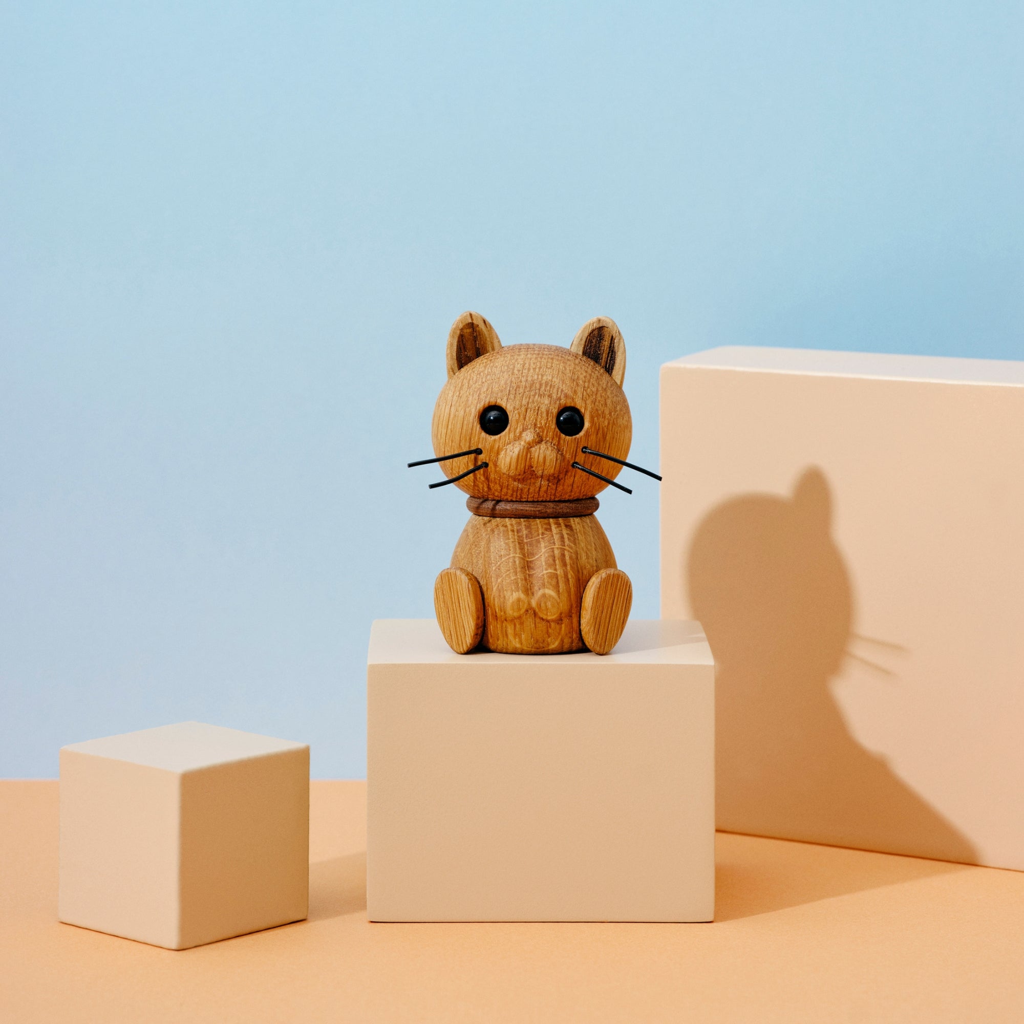 A wooden cat figurine with black whiskers sits on a beige block against a pastel blue and peach background.