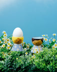 Kokeshi dolls in a field of daisies, one with an egg on its head, against a blue sky.