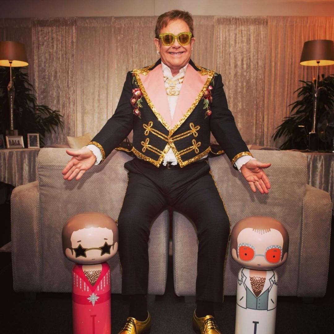 A person in a decorative outfit sits on a couch with two kokeshi dolls in front.