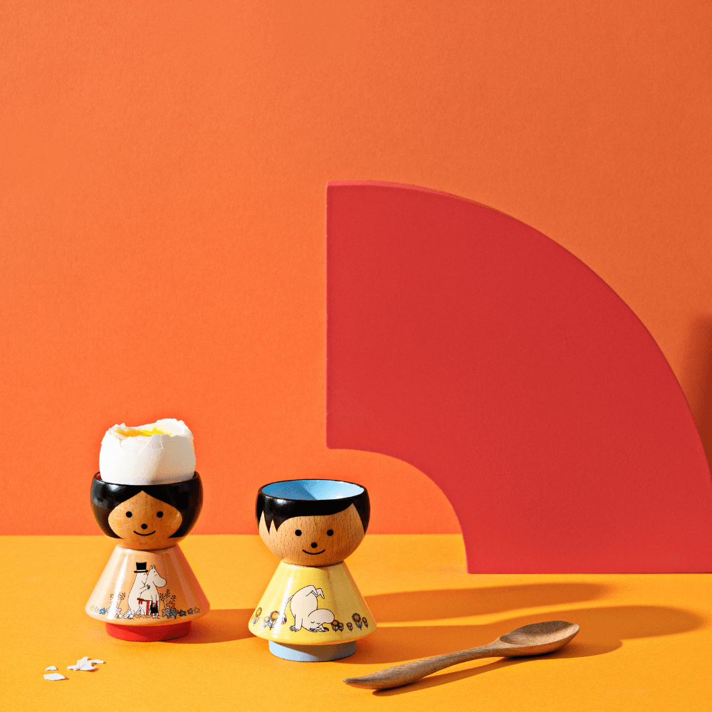 Two kokeshi doll egg cups with eggs, a wooden spoon, and a red geometric shape on an orange background.