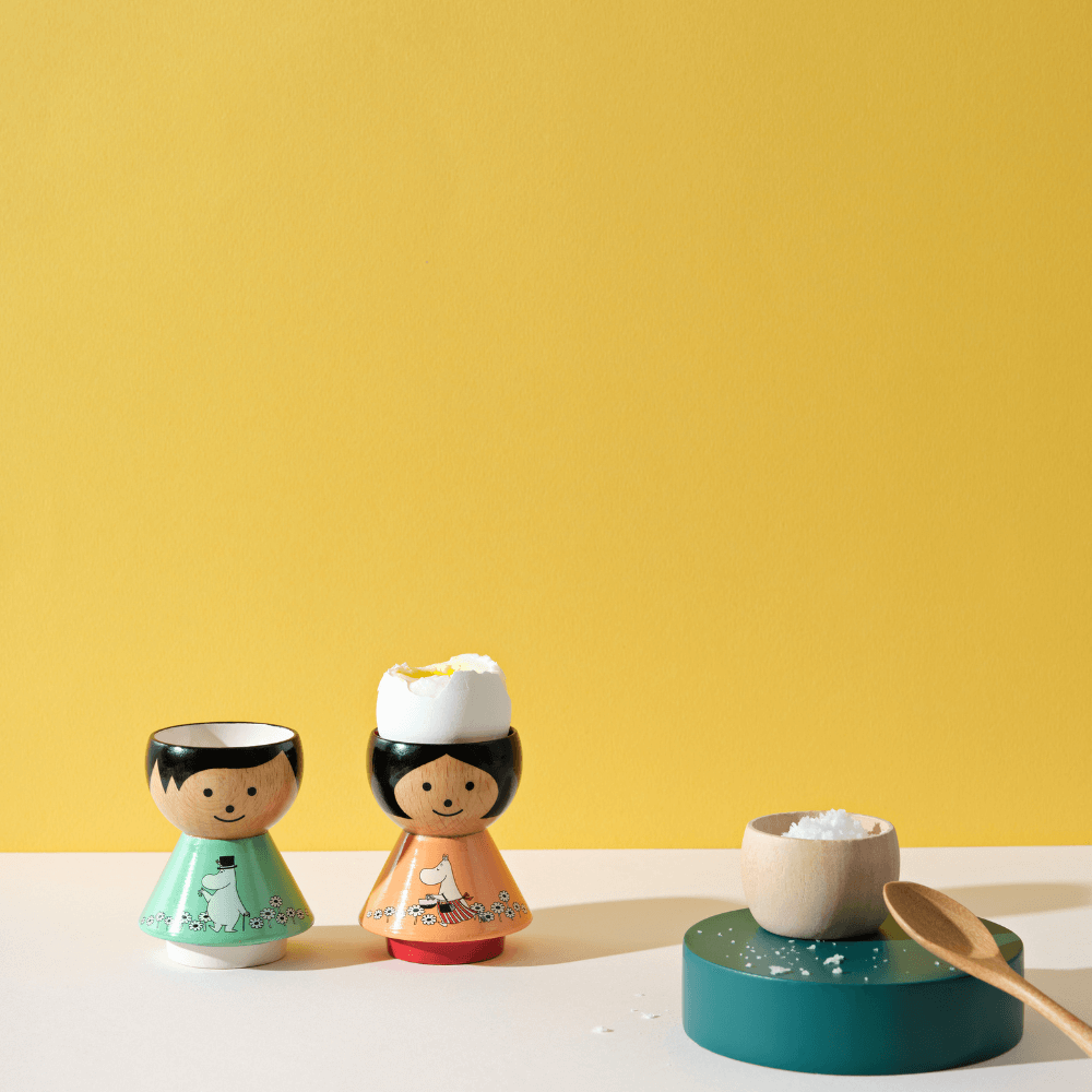 Two kokeshi doll egg cups with eggs, next to a small bowl and spoon, against a yellow background.