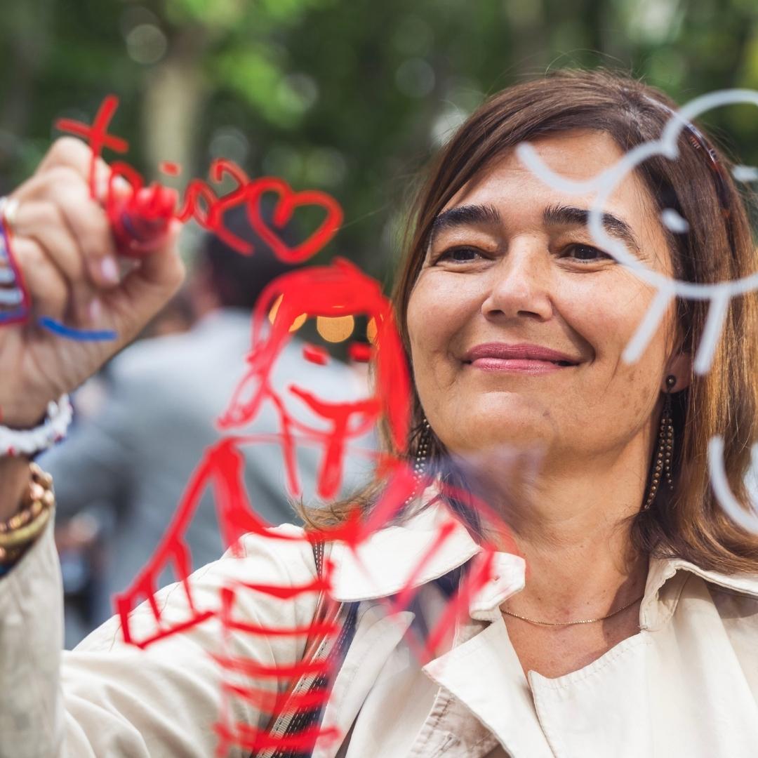 A woman drawing on a transparent surface with red and white markers, creating a figure and a sun.