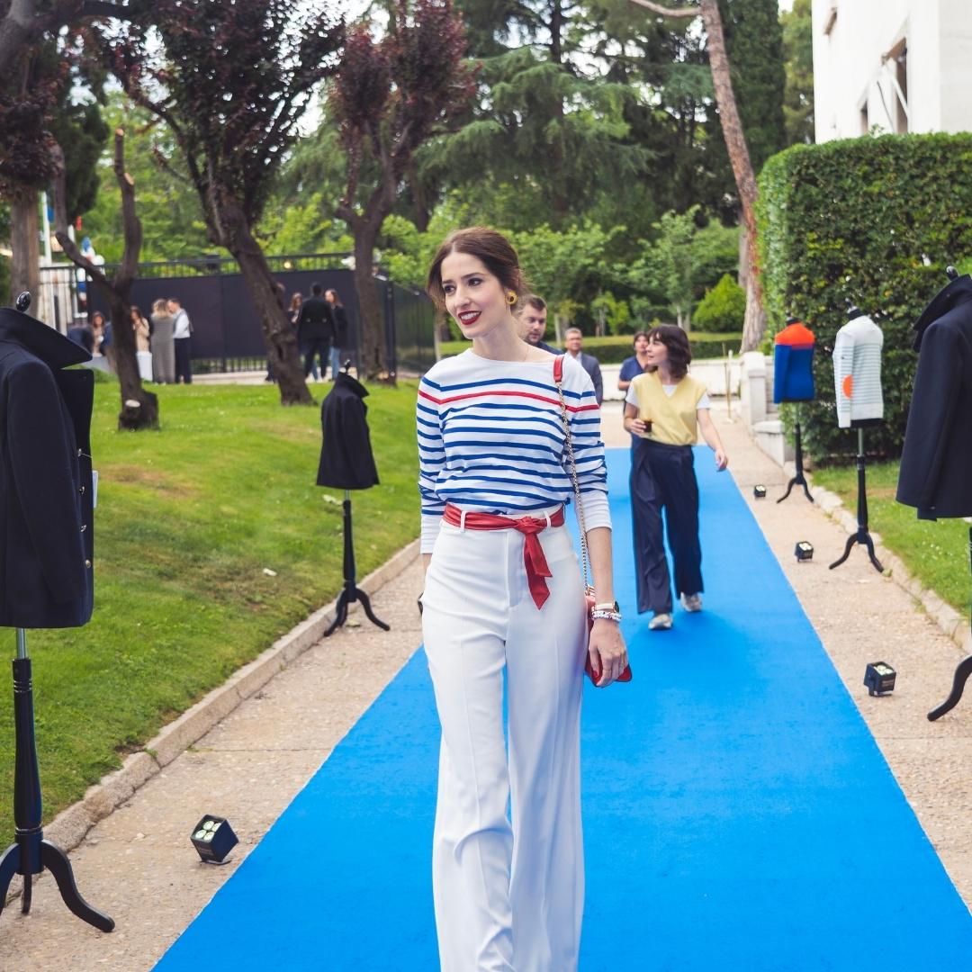 A woman in a striped shirt and white pants walks on a blue carpet outdoors, surrounded by mannequins.