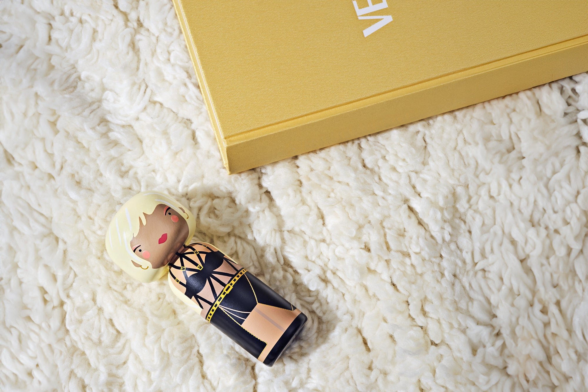 Kokeshi doll with blonde hair and black outfit on a fluffy white surface next to a yellow book.