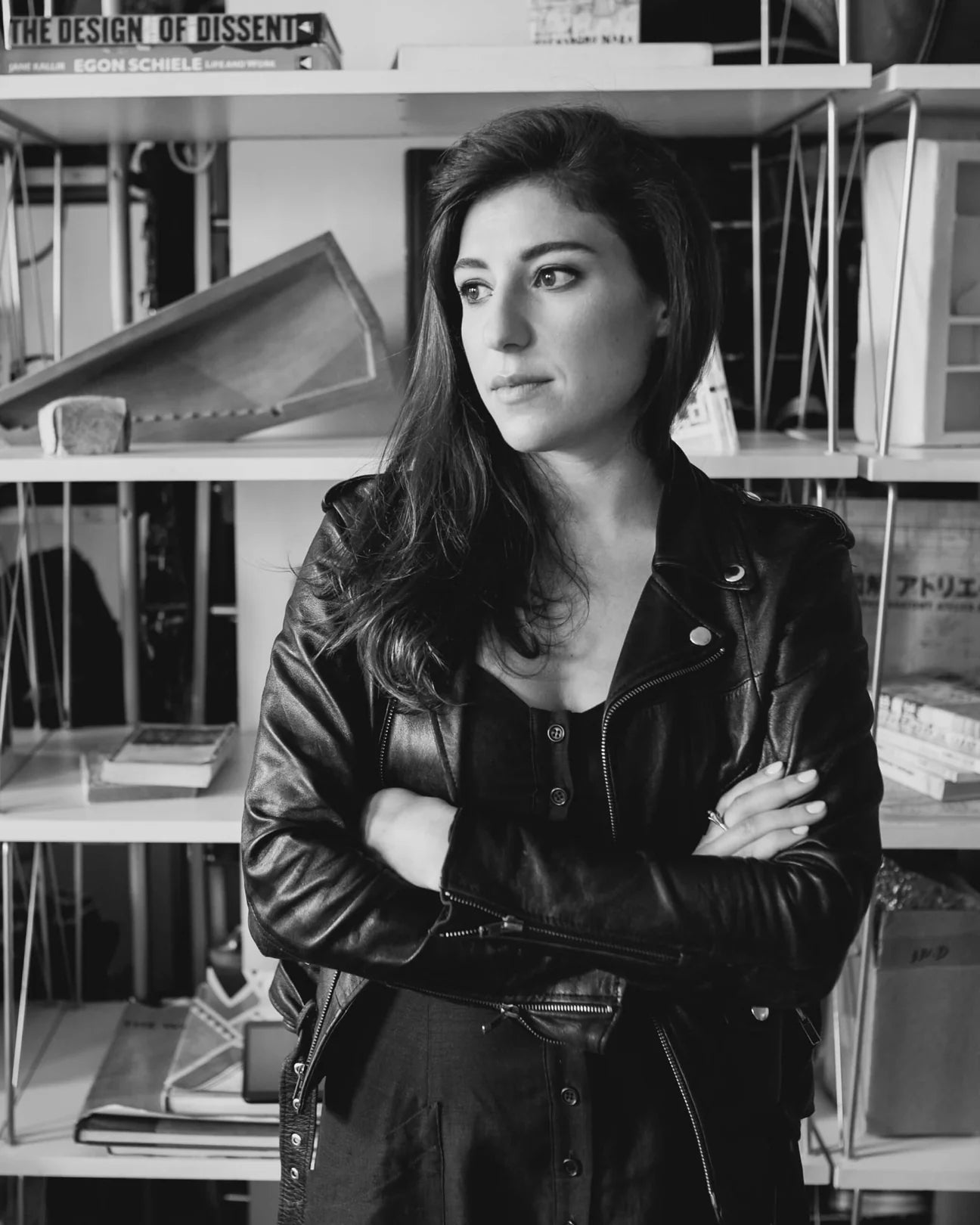 A person in a leather jacket stands with arms crossed in front of shelves filled with books and decor items.