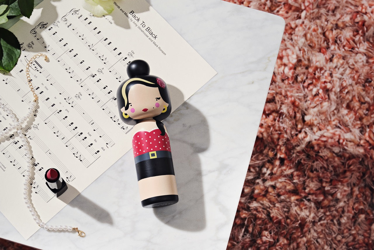 Kokeshi doll on a marble table with sheet music, lipstick, and a pearl necklace nearby.