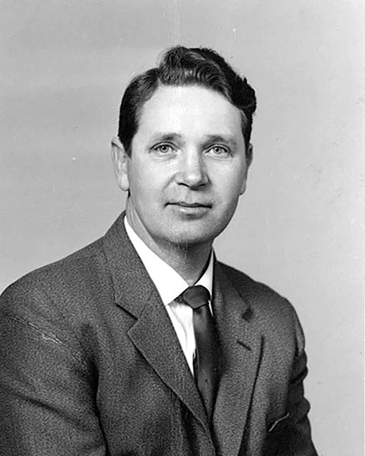 A black and white portrait of a man in a suit and tie, with short hair, looking at the camera.