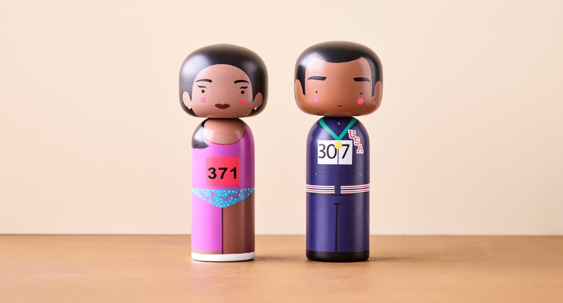 Two kokeshi dolls: one in a pink swimsuit with number 371, the other in a blue tracksuit with number 307.