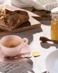 A pink teacup with tea, a spoon, a loaf of bread, honey jar, and newspaper on a table.