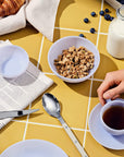 A breakfast scene with cereal, tea, a croissant, blueberries, and a newspaper on a yellow tiled table.
