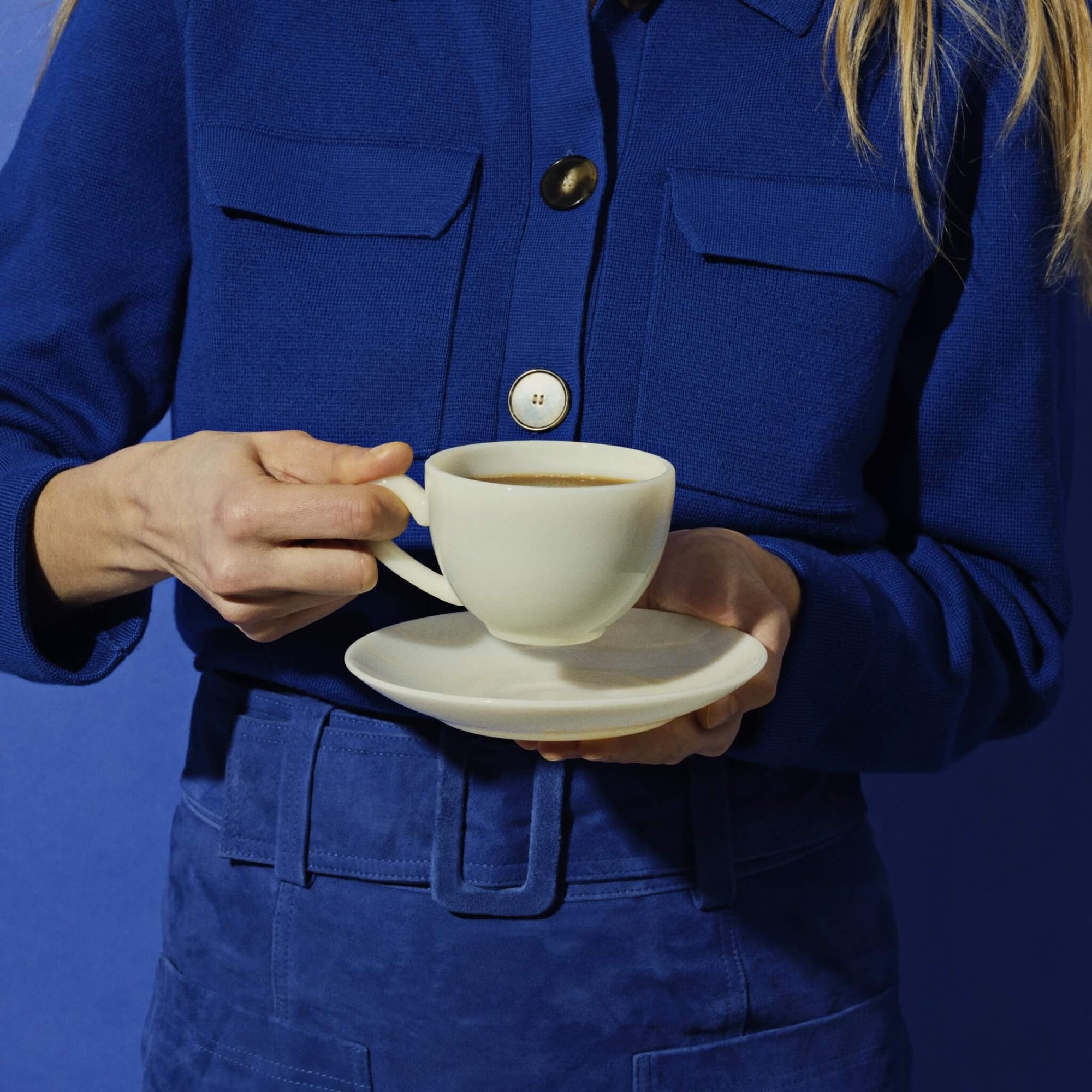 A person in a blue outfit holding a white cup and saucer against a blue background.