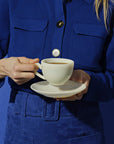 A person in a blue outfit holding a white cup and saucer against a blue background.