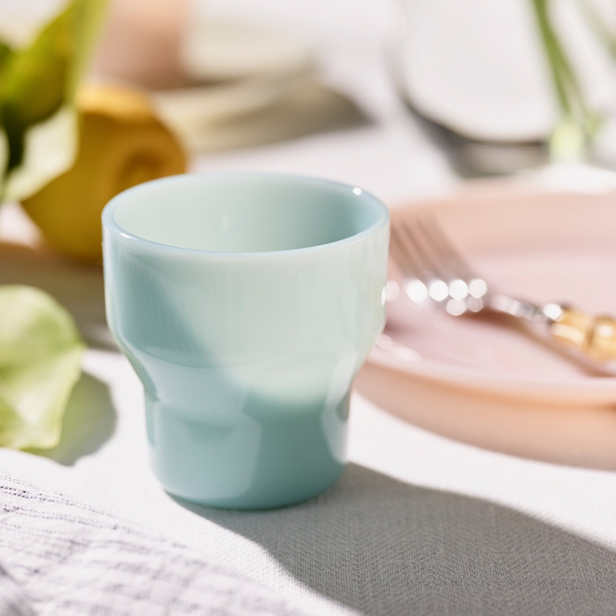 A light blue ceramic cup on a table with a pink plate, fork, and blurred background elements.
