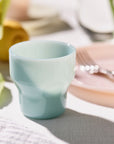 A light blue ceramic cup on a table with a pink plate, fork, and blurred background elements.