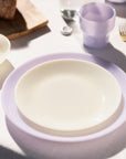 A table setting with a white plate on a lavender plate, next to a purple cup and gold cutlery.
