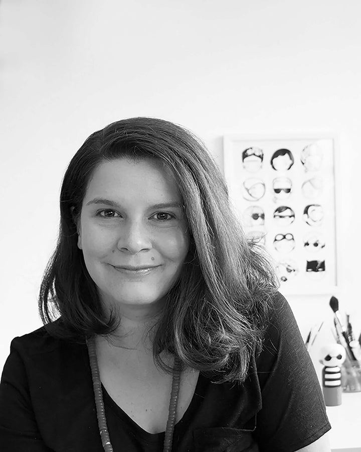 A person with shoulder-length hair smiling, with artwork and brushes in the background.