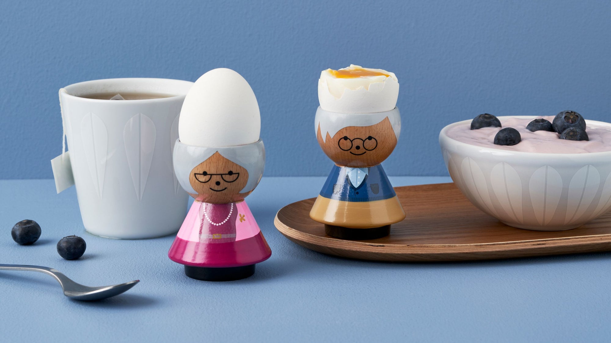 Kokeshi doll egg cups with eggs, a cup of tea, and a bowl of yogurt with blueberries on a blue surface.