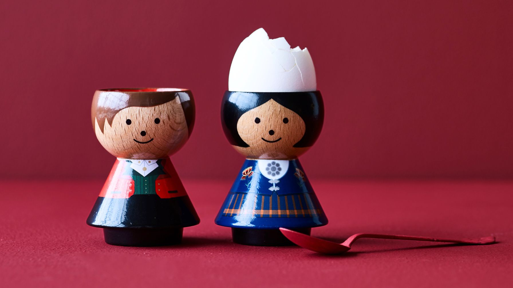 Two kokeshi doll egg cups with smiling faces, one holding a cracked egg, on a red background.