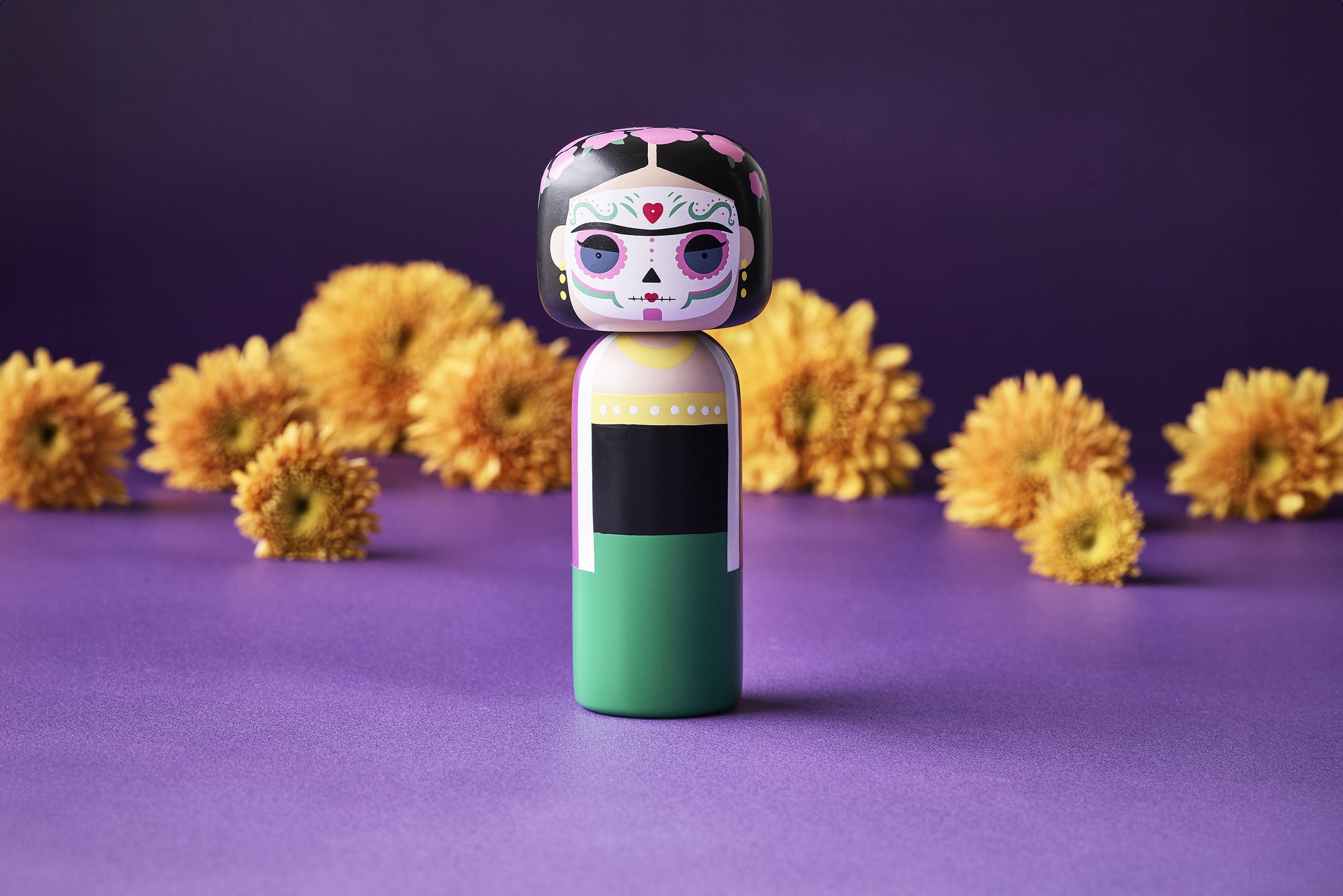 A kokeshi doll with a colorful design stands on a purple surface, surrounded by yellow flowers.