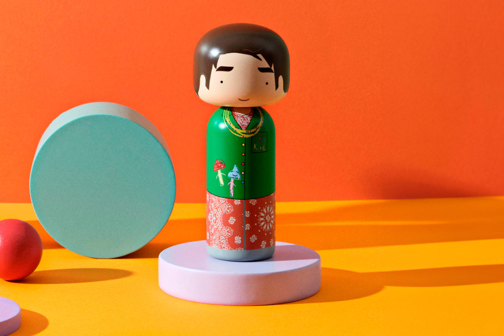 A kokeshi doll with a green top and red patterned skirt on a pastel platform against an orange background.