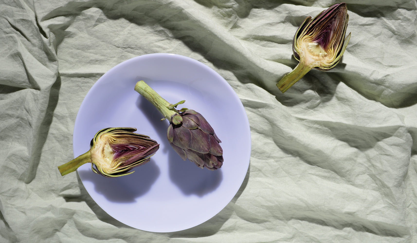 A whole artichoke and two halves on a white plate and green fabric background.