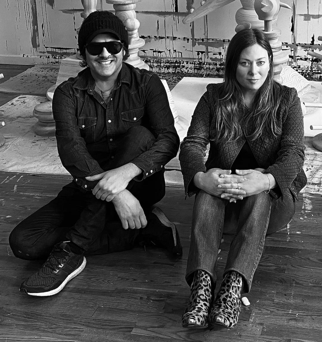 Two people sitting on the floor in a room with large wooden objects in the background.