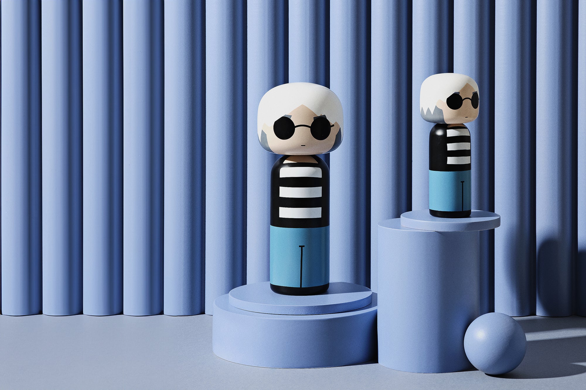 Two kokeshi dolls with striped shirts and sunglasses on blue cylindrical platforms against a striped backdrop.