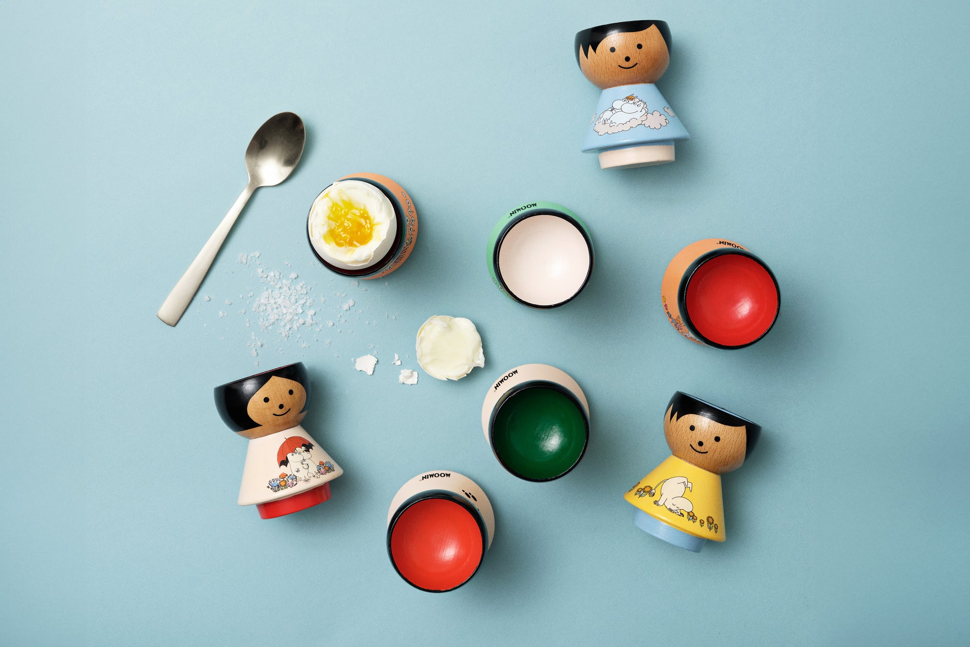 Kokeshi doll egg cups with colorful interiors and a spoon on a blue background.