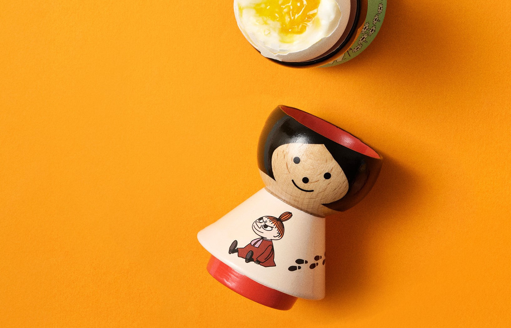 A kokeshi doll with a smiling face and a red outfit on an orange background, next to a soft-boiled egg.