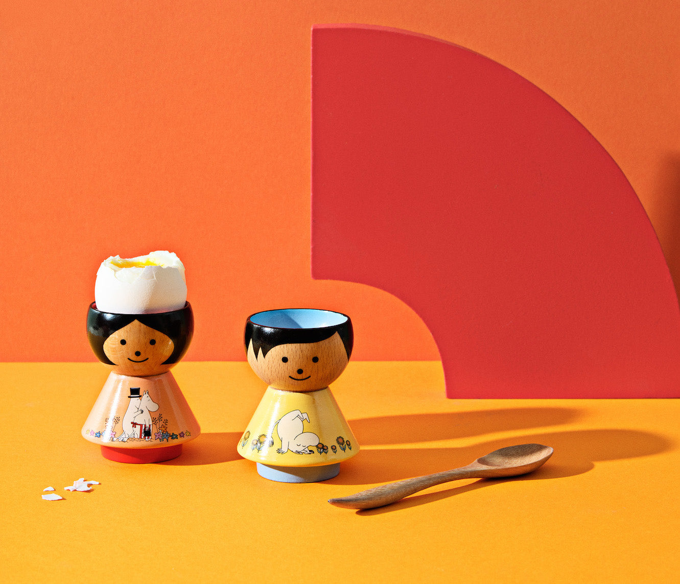 Two kokeshi doll egg cups with painted designs, one holding an egg, on an orange and red background.