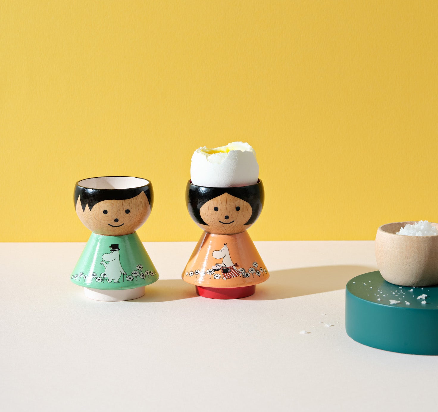 Two kokeshi doll egg cups, one with a cracked egg, on a table with a salt bowl and yellow background.
