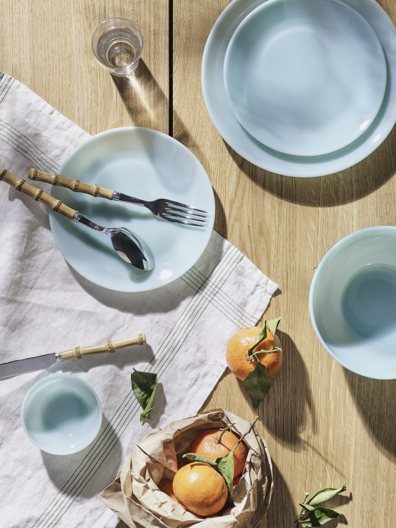 Light blue plates and bowls on a wooden table with oranges and cutlery on a striped cloth.