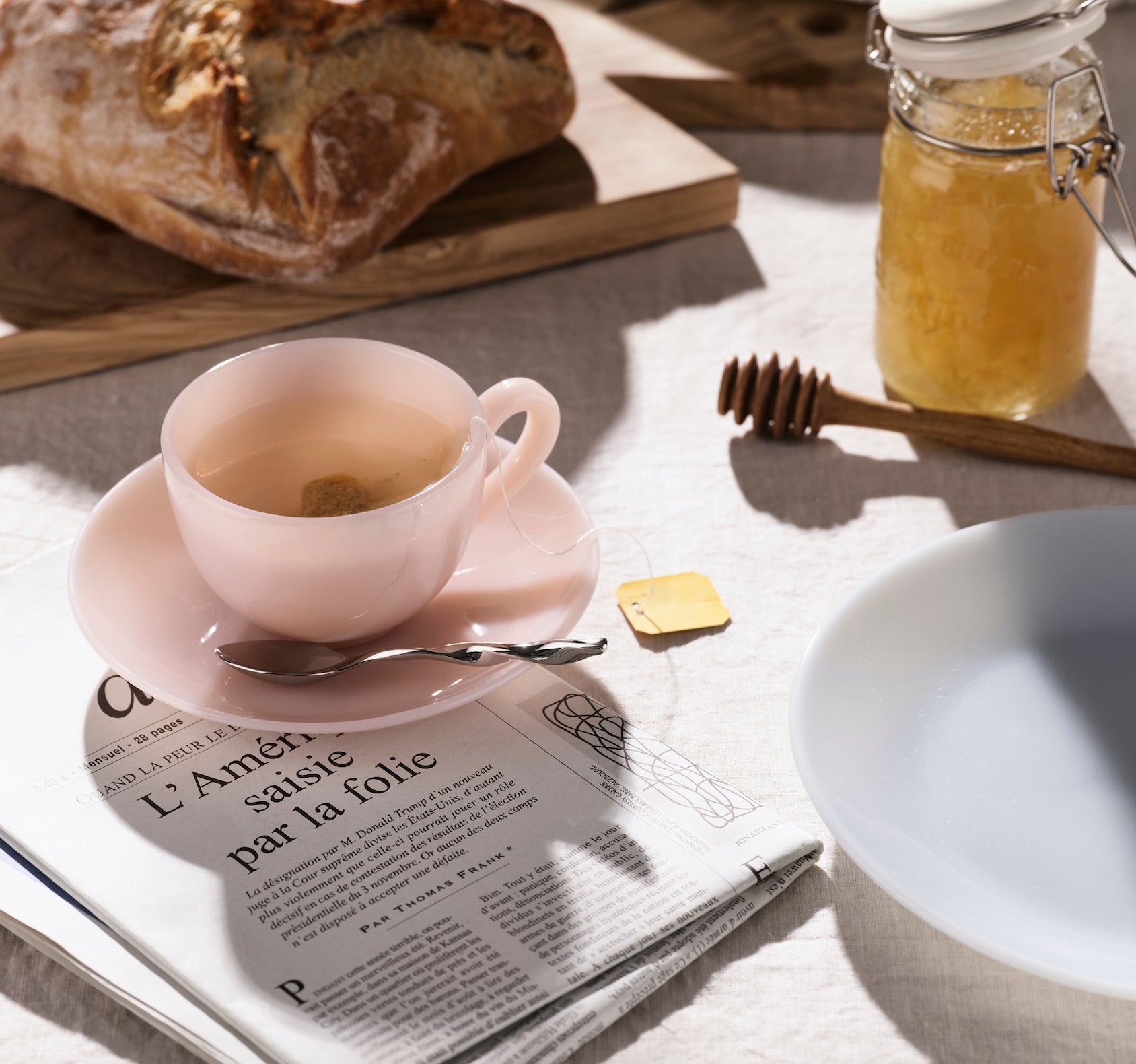 A pink teacup on a newspaper with bread, honey, and a honey dipper on a sunlit table.