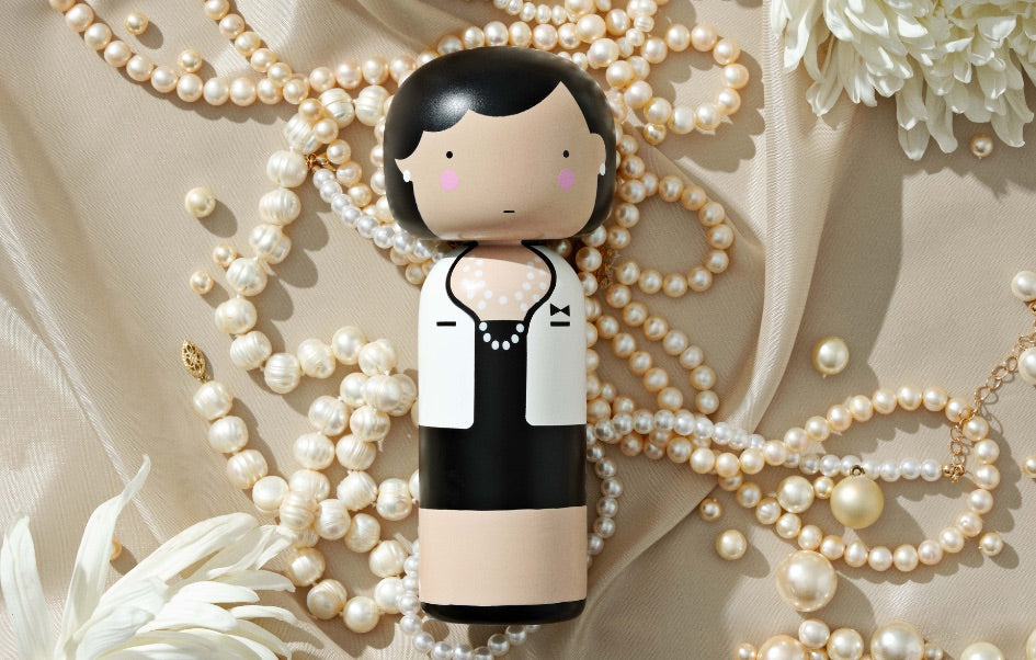 A kokeshi doll with a black and white outfit surrounded by pearl necklaces and white flowers.