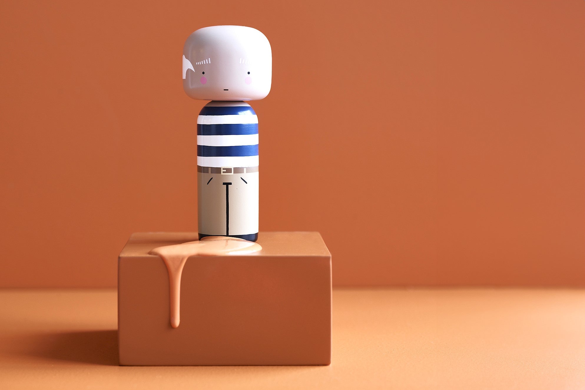 A kokeshi doll with a striped shirt stands on a brown block with a paint drip on an orange background.