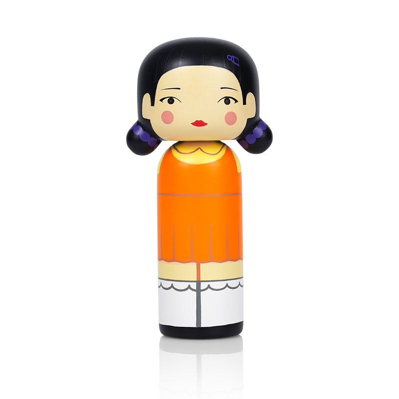 A kokeshi doll with black hair, orange dress, and white shoes, featuring a simple, stylized design.