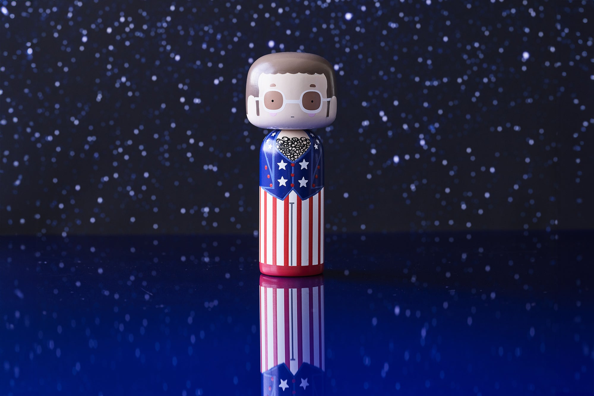 A kokeshi doll with a starry background, wearing a suit with stars and stripes, reflecting on a blue surface.
