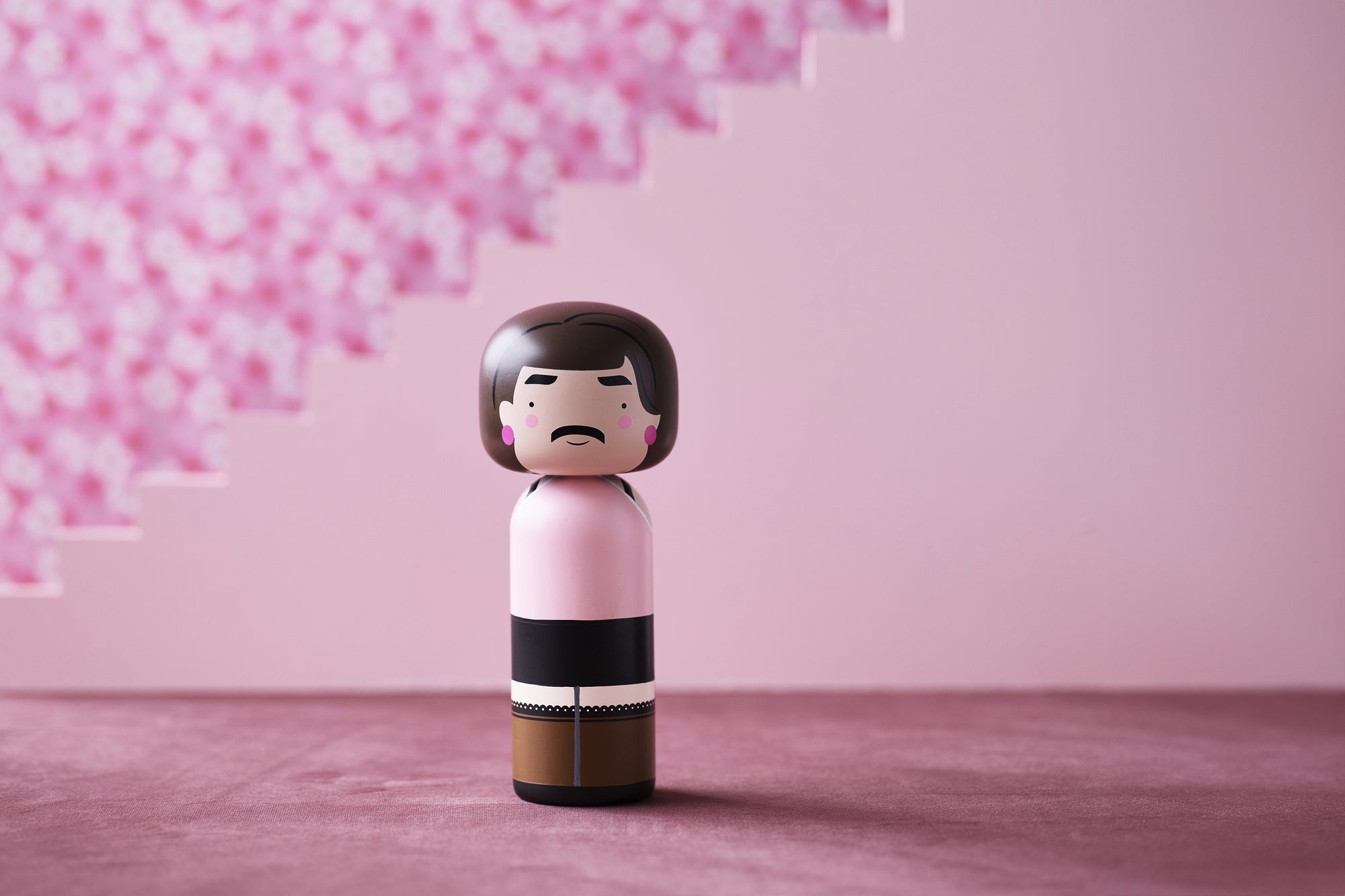 A kokeshi doll with a pink and brown outfit stands on a pink surface with a pink floral background.