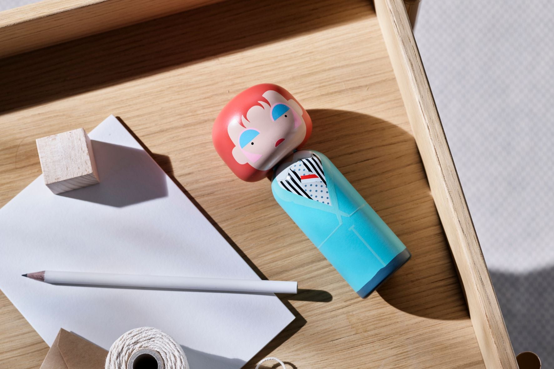 Kokeshi doll in a blue suit on a wooden tray with a pencil, paper, and twine.