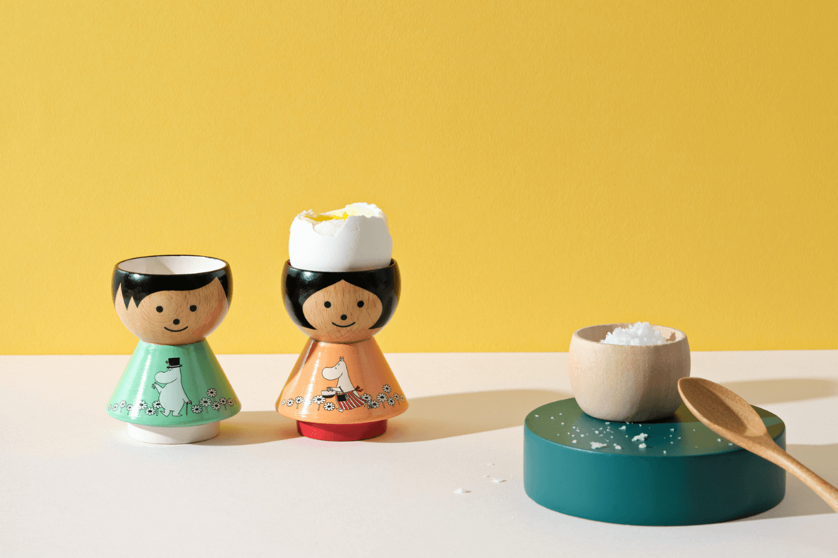 Two kokeshi egg cups with painted designs, one holding an egg, next to a small bowl of salt and a wooden spoon.