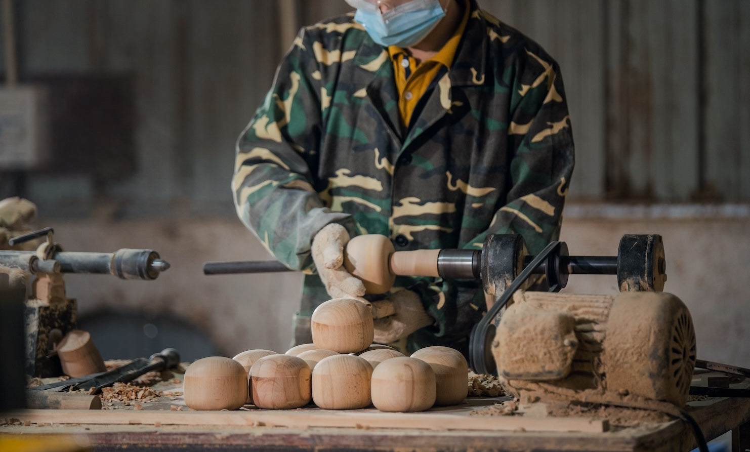 A person in a camo jacket and mask crafting wooden dolls on a lathe in a workshop.