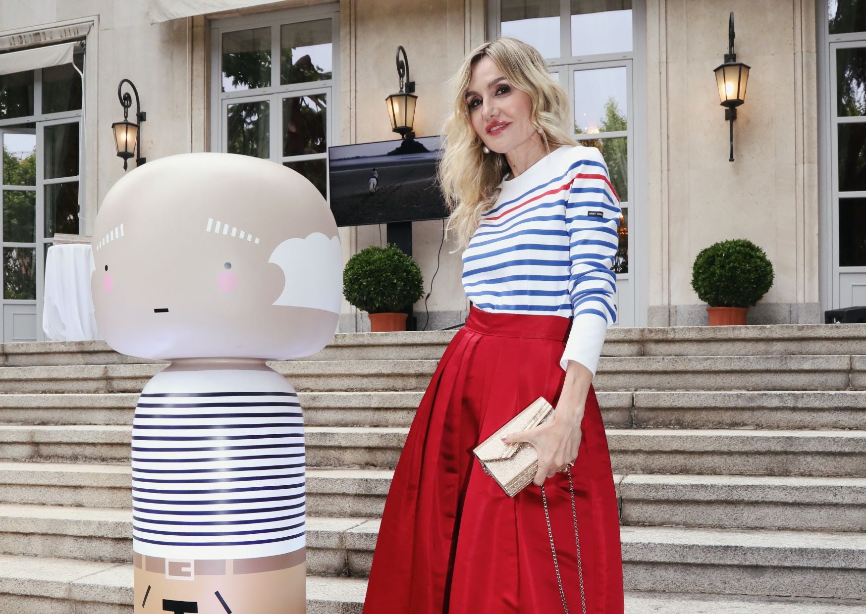 A woman in a striped shirt and red skirt stands beside a large kokeshi doll outdoors.