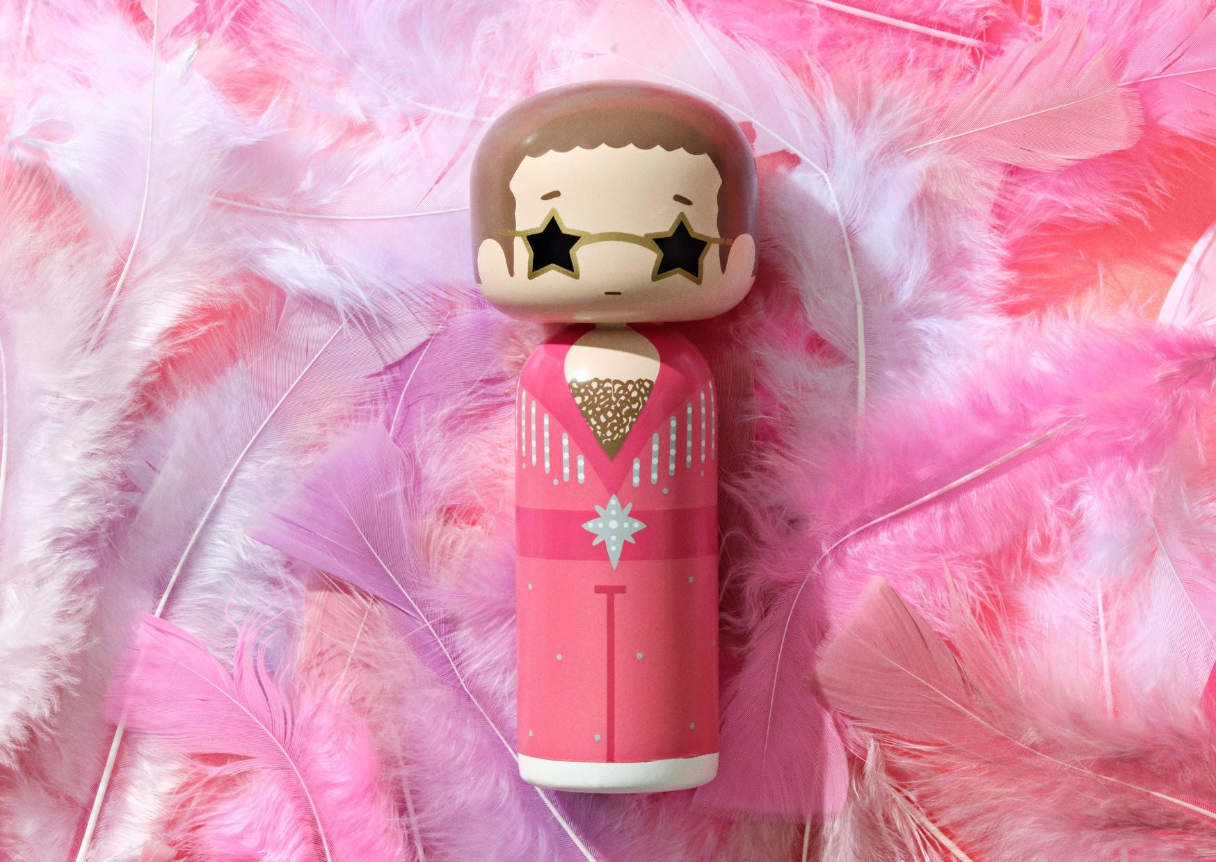 A kokeshi doll in a pink outfit with star-shaped sunglasses, surrounded by pink and white feathers.