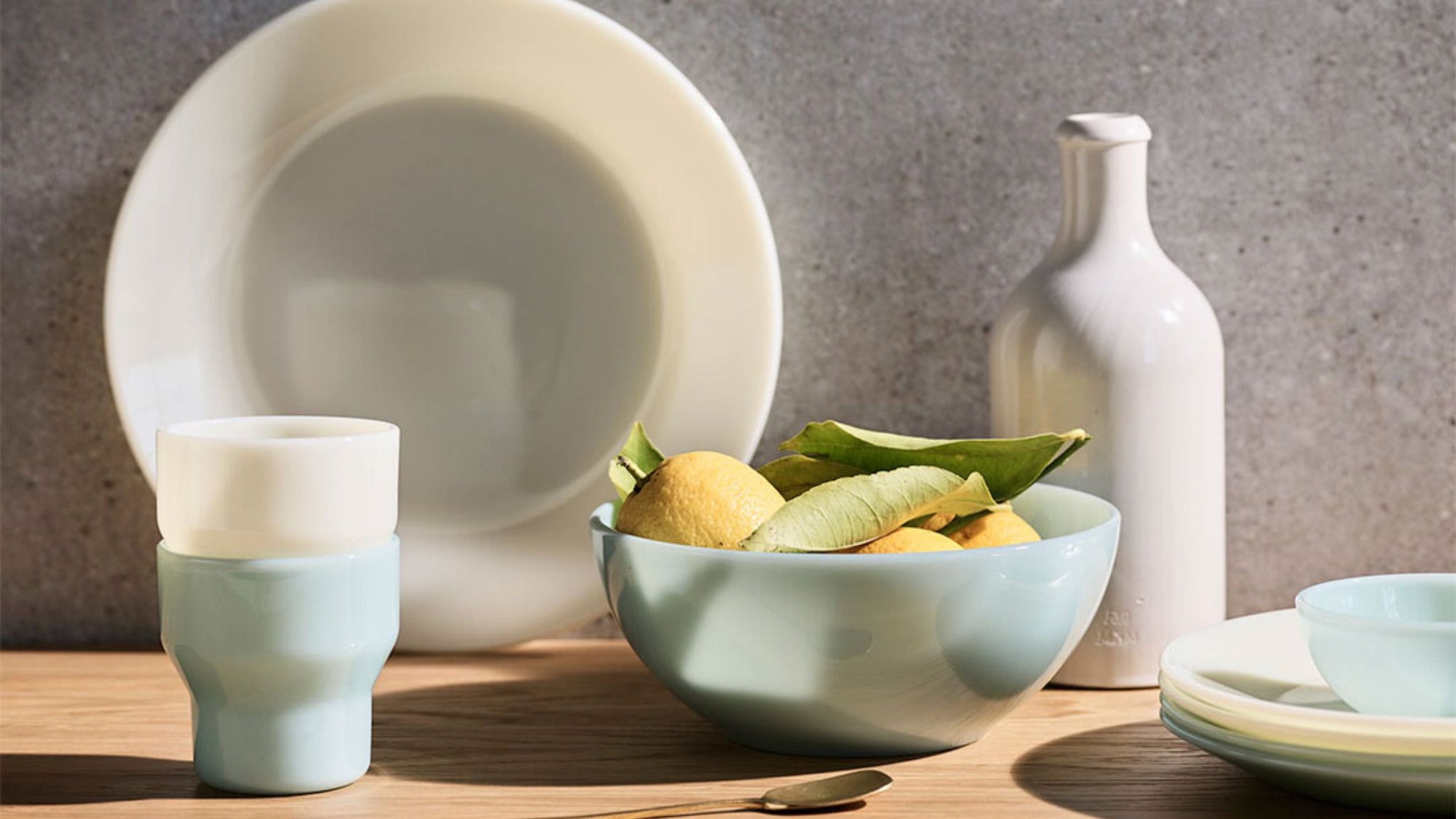 Ceramic dishes, a bowl of lemons, and a bottle on a wooden table against a gray wall.