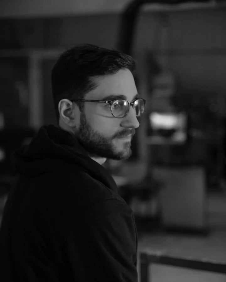 A person with glasses and a beard in a dimly lit room, looking to the side.