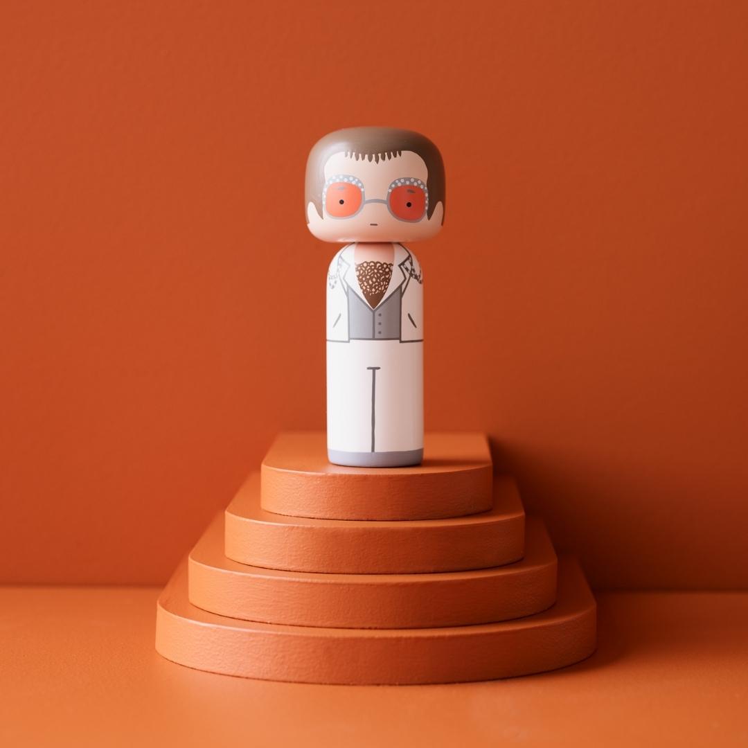 En kokeshi-dukke med briller og jakkesæt på en orange platform mod en orange baggrund.