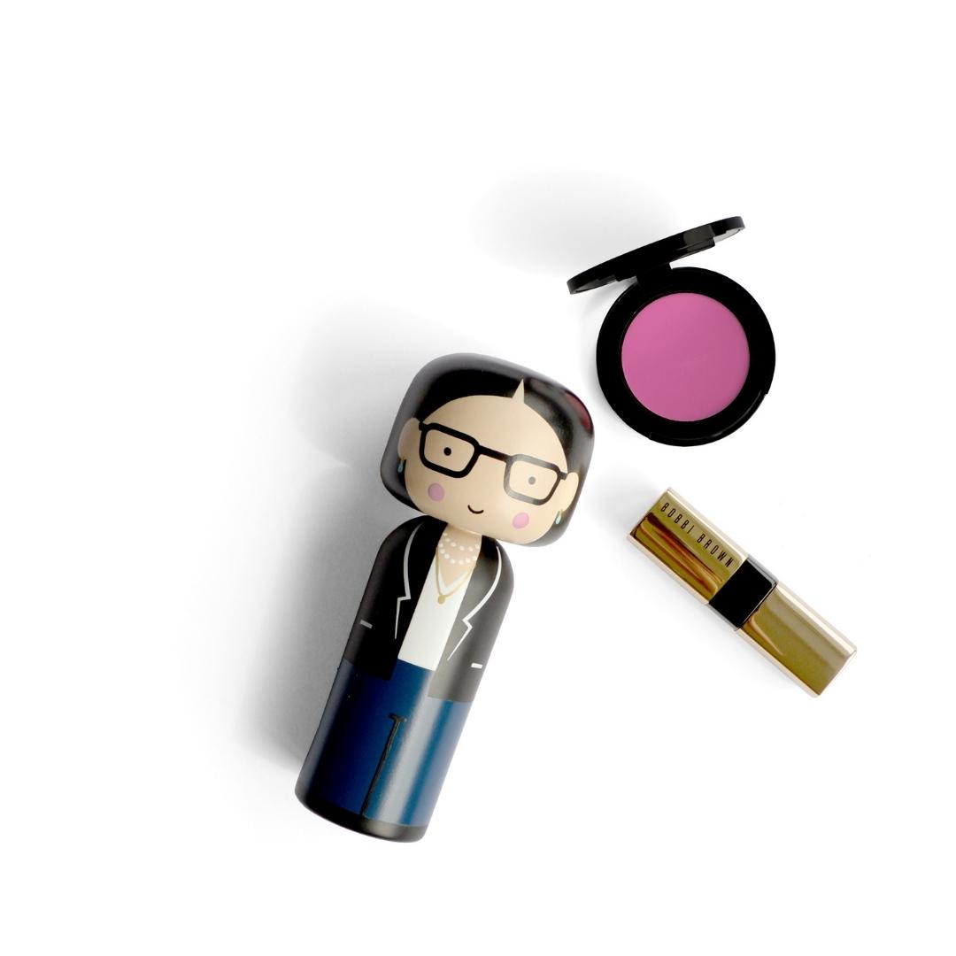 Kokeshi-dukke med briller, sort blazer og blå bukser ved siden af pink blush og guldlæbestift.